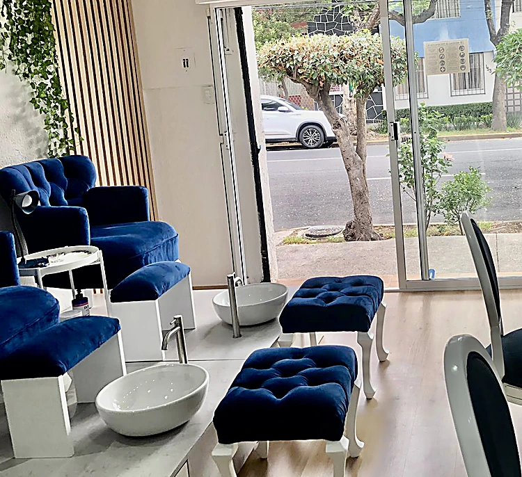 Interior del salón: sillones de pedicure capitoné azul marino, lavabos blancos, taburetes de terciopelo y un ventanal hacia la calle con árboles